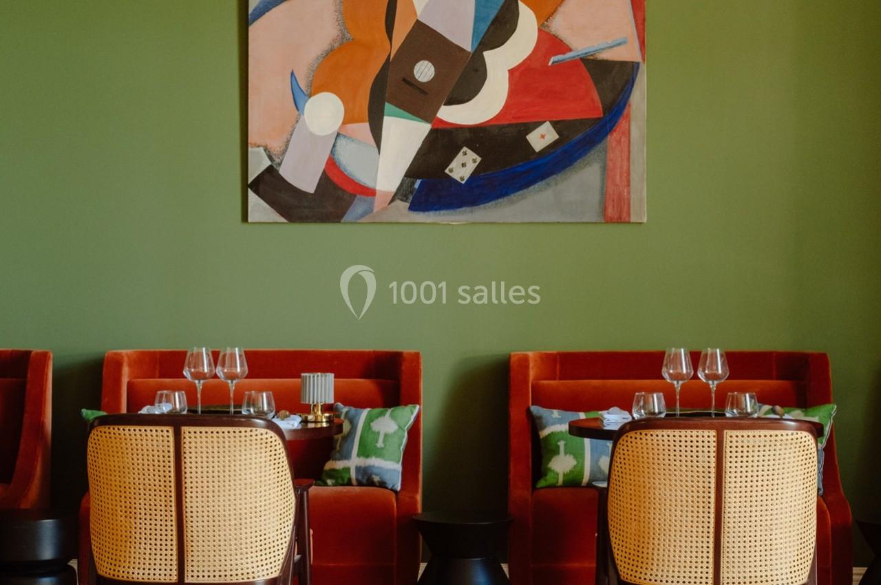Salle de restaurant avec murs verts, tables dressées, chaises en rotin et tableau abstrait coloré au mur.