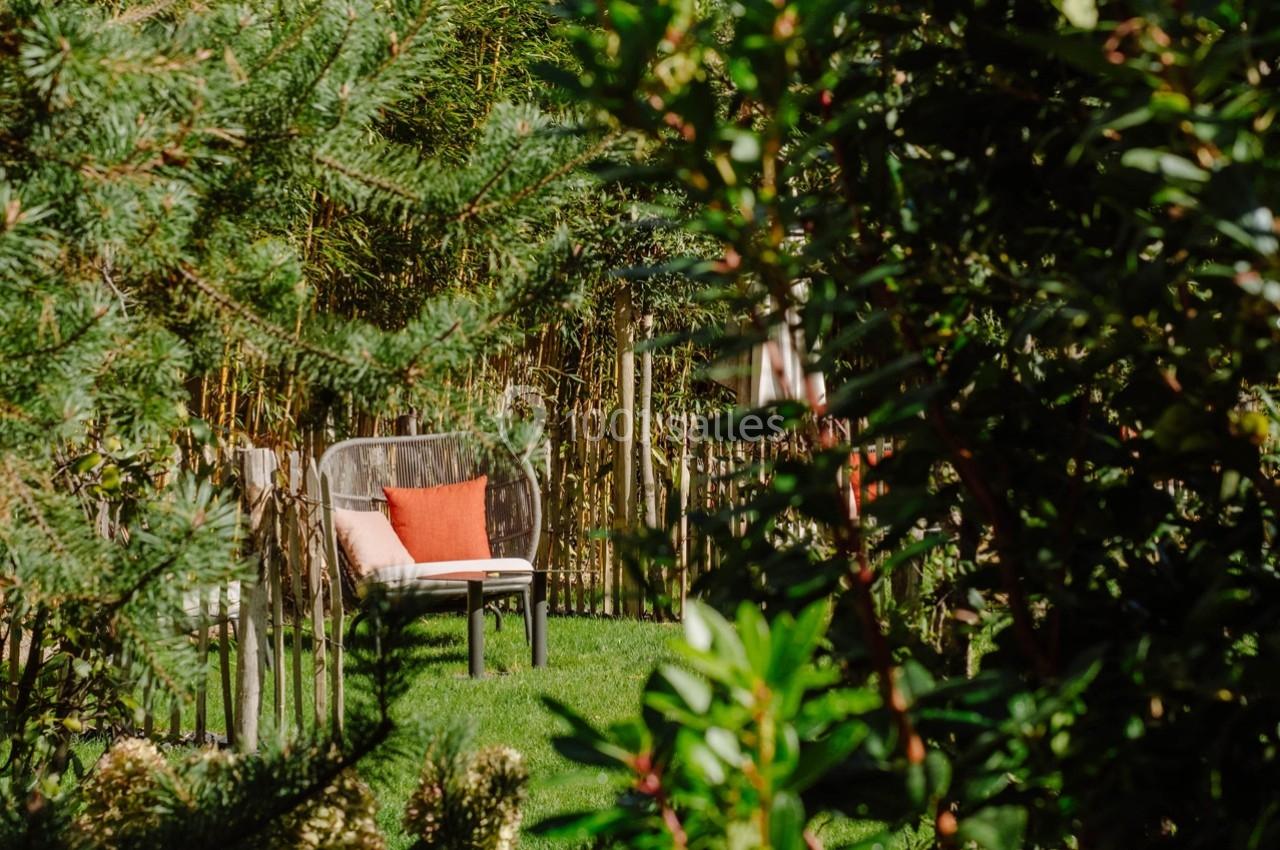 Banc en métal avec coussin orange, placé dans un jardin verdoyant entouré de plantes et d'arbustes.