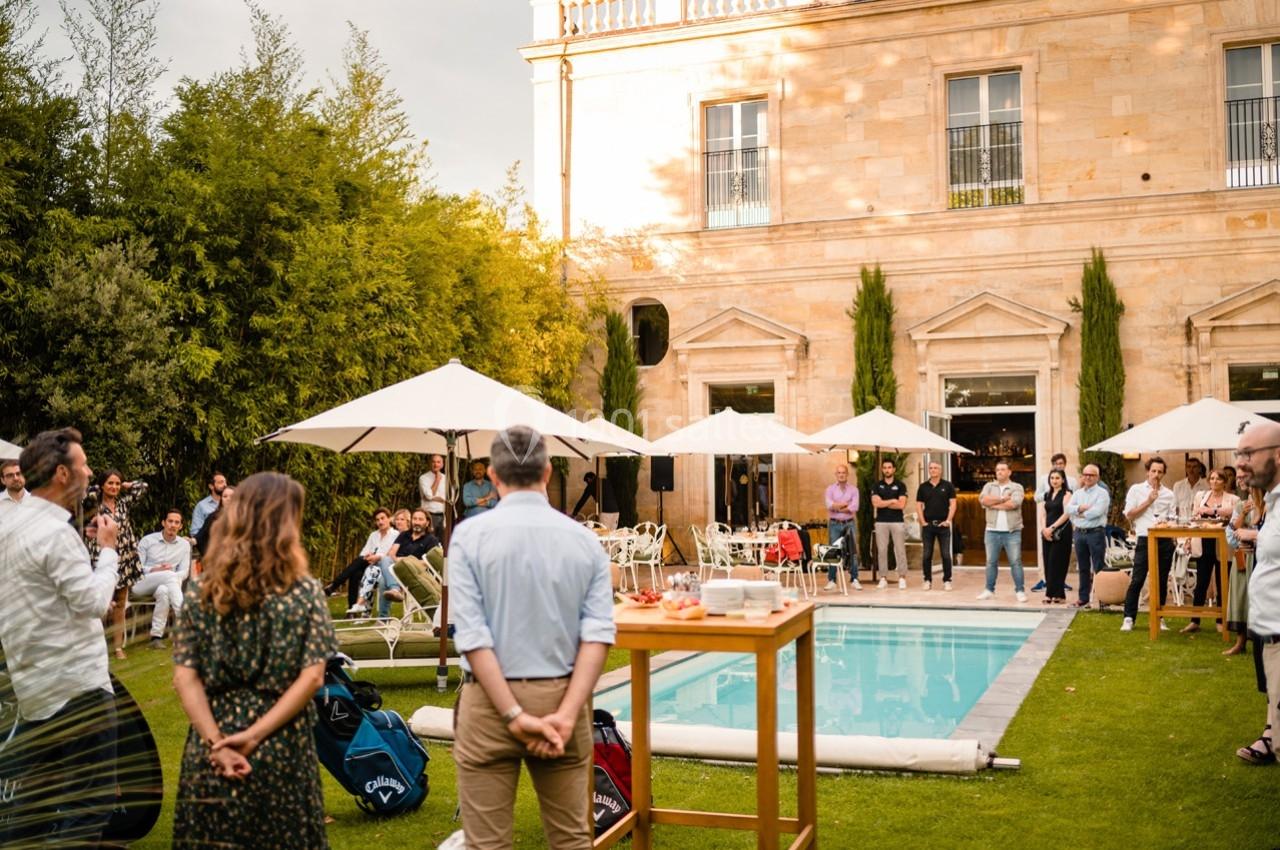 Groupe de personnes rassemblées autour d'une piscine dans un jardin, devant un bâtiment en pierre avec des parasols.