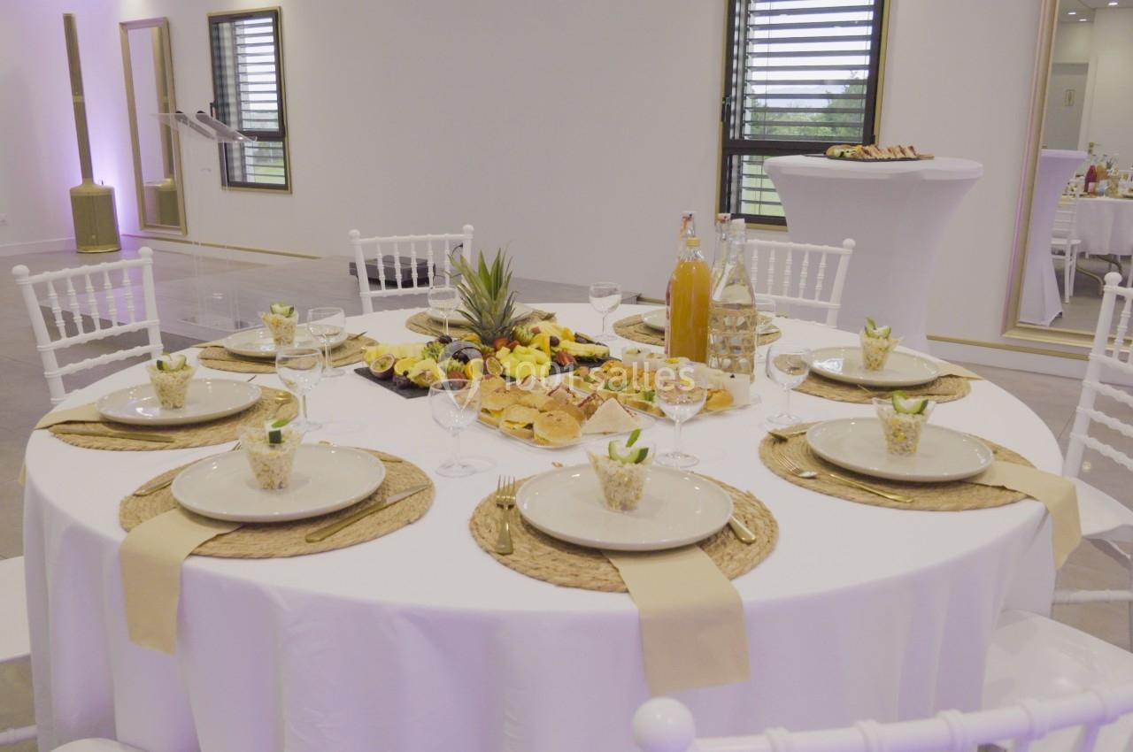 Table ronde dressée avec nappes blanches, assiettes, verres, décorations et plats variés dans une salle lumineuse.