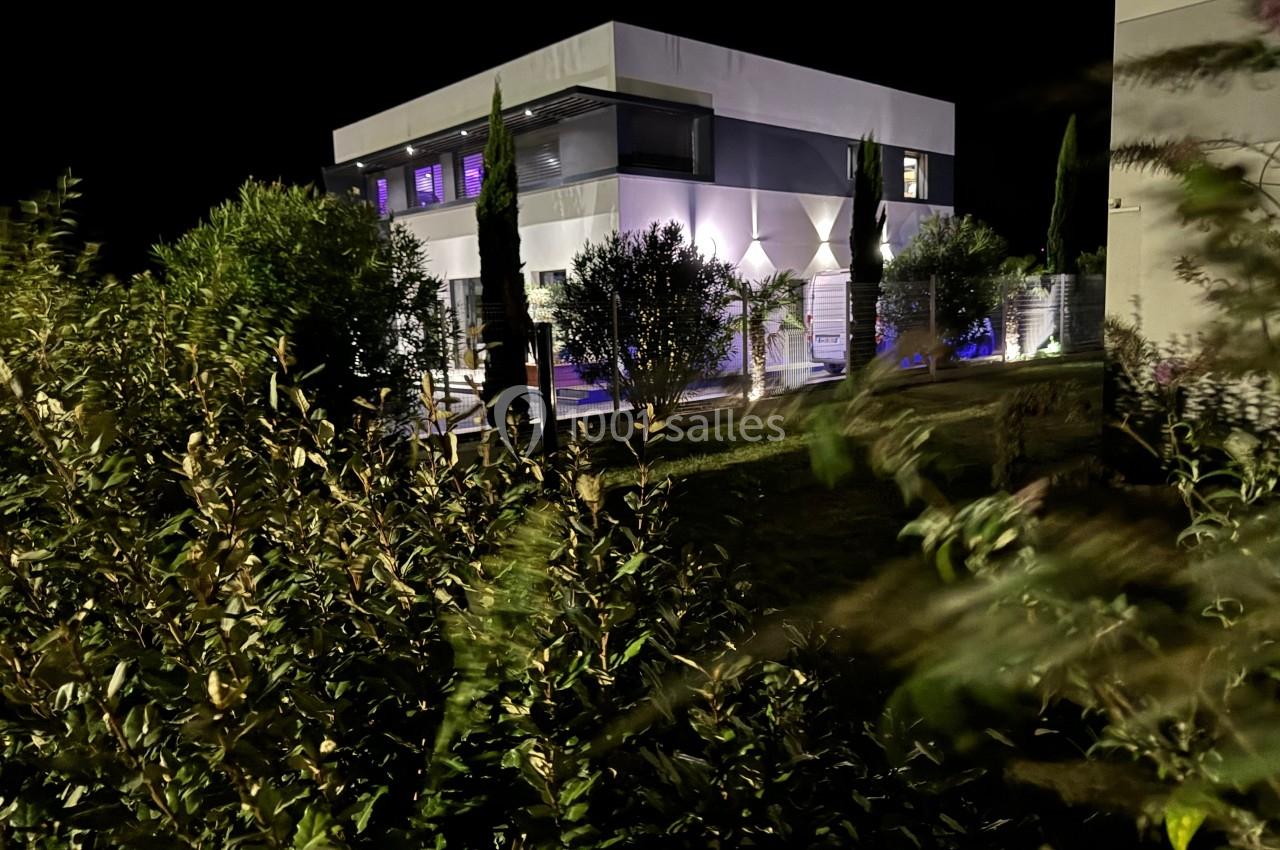 Maison moderne éclairée la nuit, entourée de végétation et d'arbustes au premier plan.