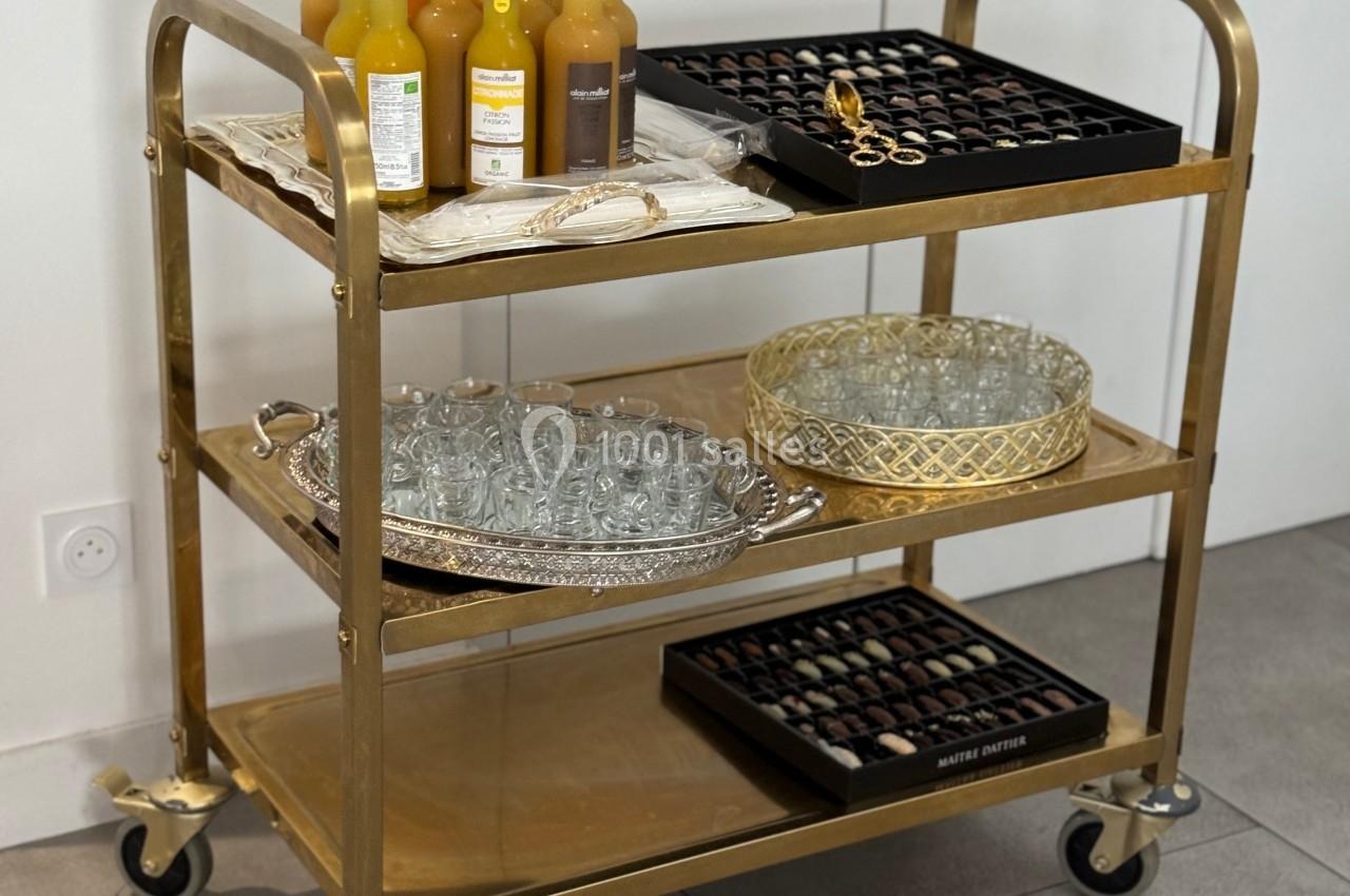 Chariot doré à trois étagères contenant des bouteilles de jus, des verres, un plateau et des chocolats.
