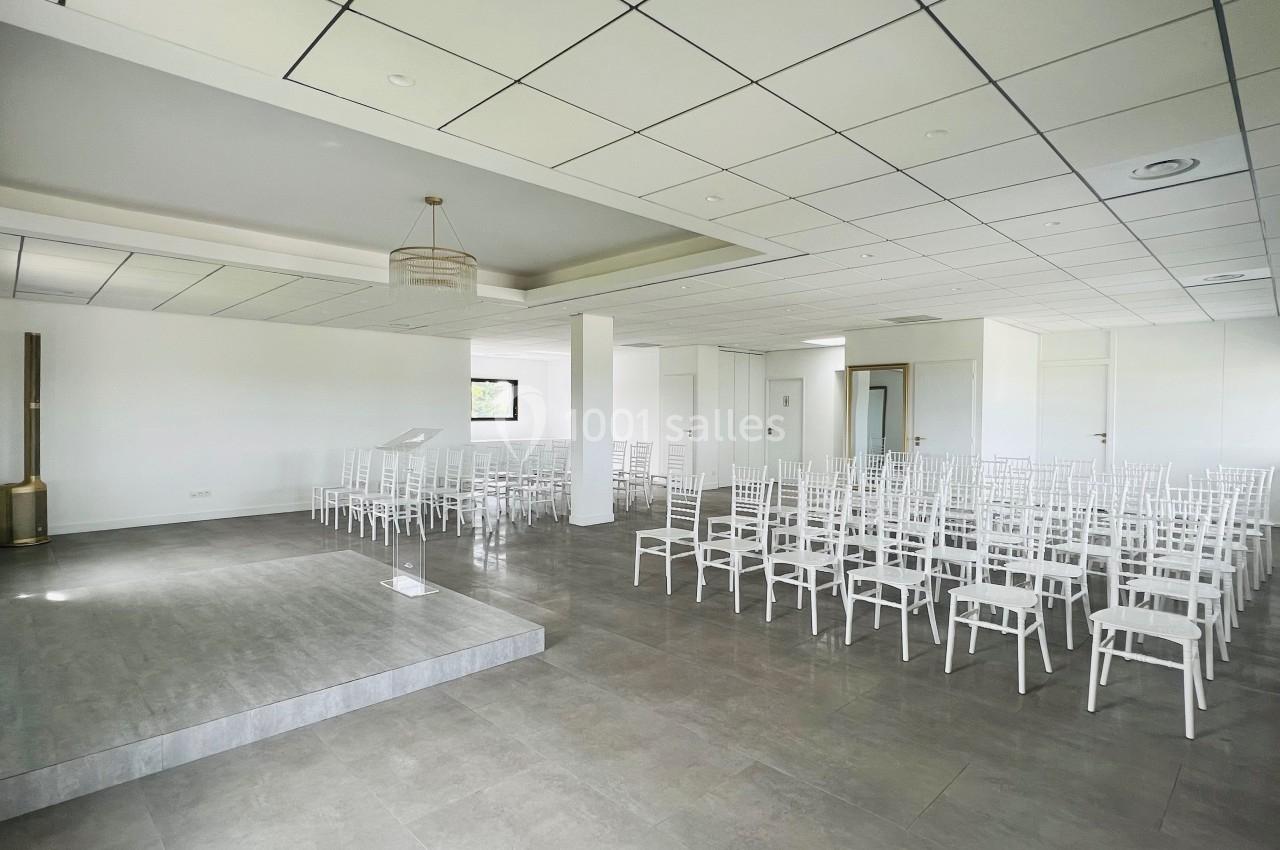Salle lumineuse avec des chaises blanches alignées face à une petite estrade sur un sol en carrelage gris.