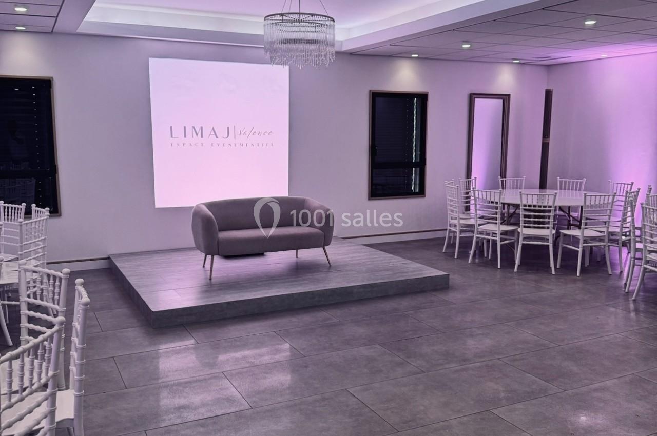 Salle de réception moderne avec éclairage violet, chaises blanches, estrade centrale et canapé gris.