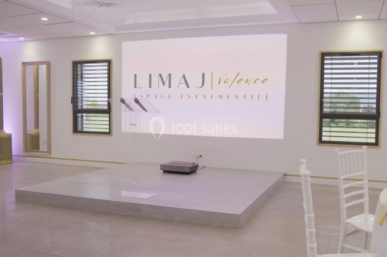 Salle lumineuse avec estrade, écran de projection, chaises blanches et fenêtres à persiennes donnant sur un extérieur…