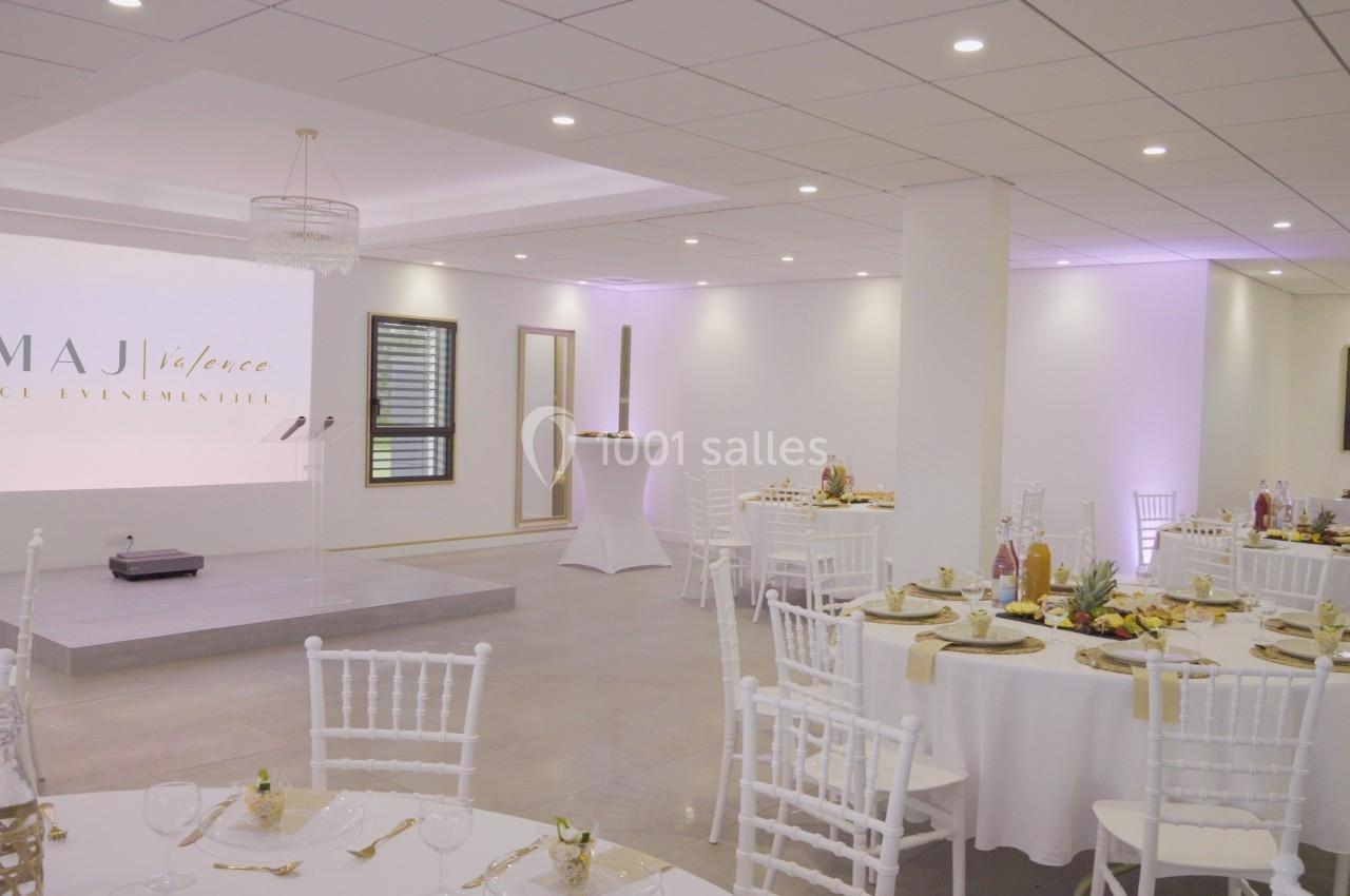 Salle de réception lumineuse avec tables rondes décorées, chaises blanches et une scène en arrière-plan.