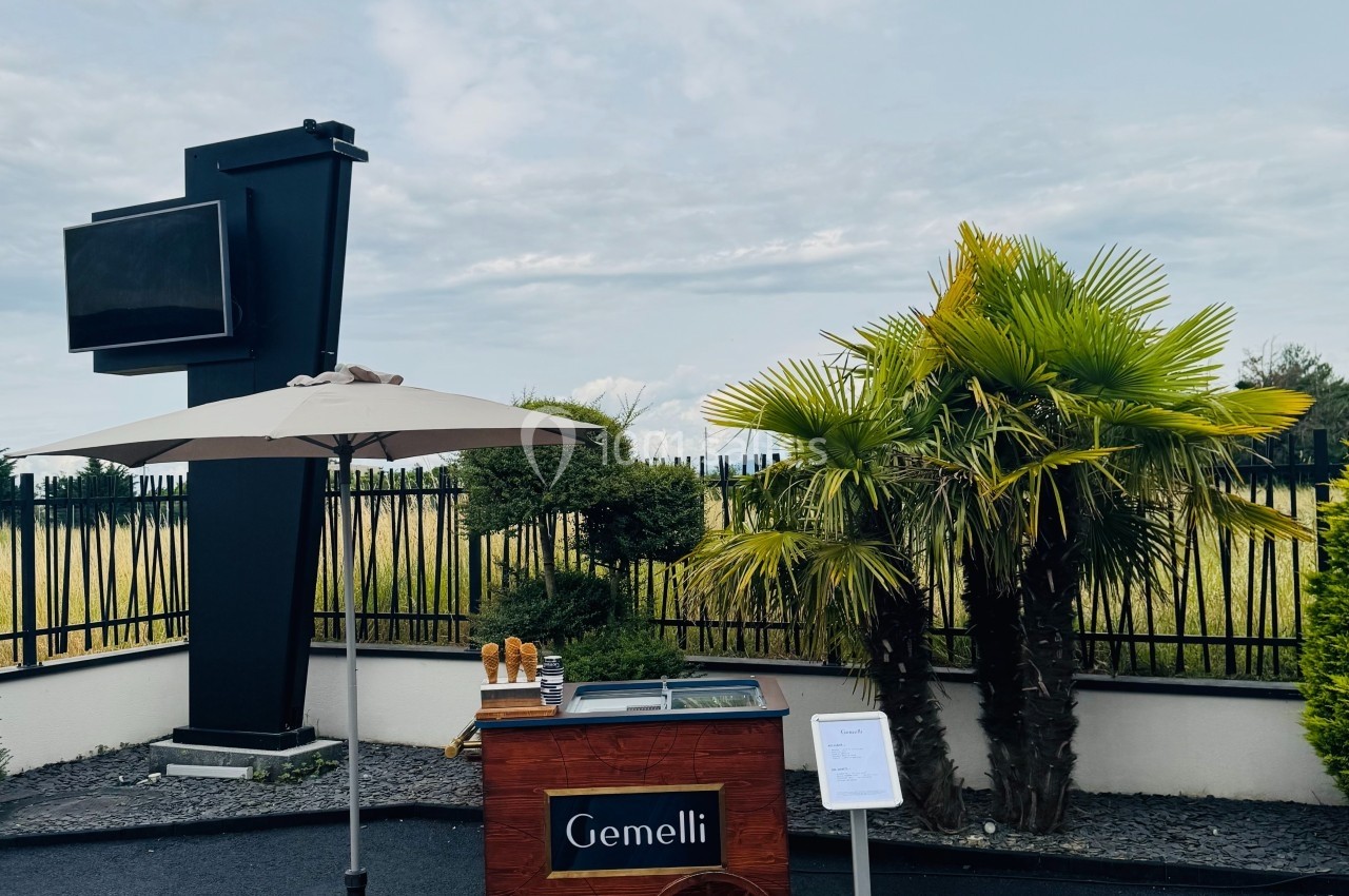 Chariot en bois avec parasol, marqué ’Gemelli’, disposé sur un parking près de palmiers et d'une clôture.