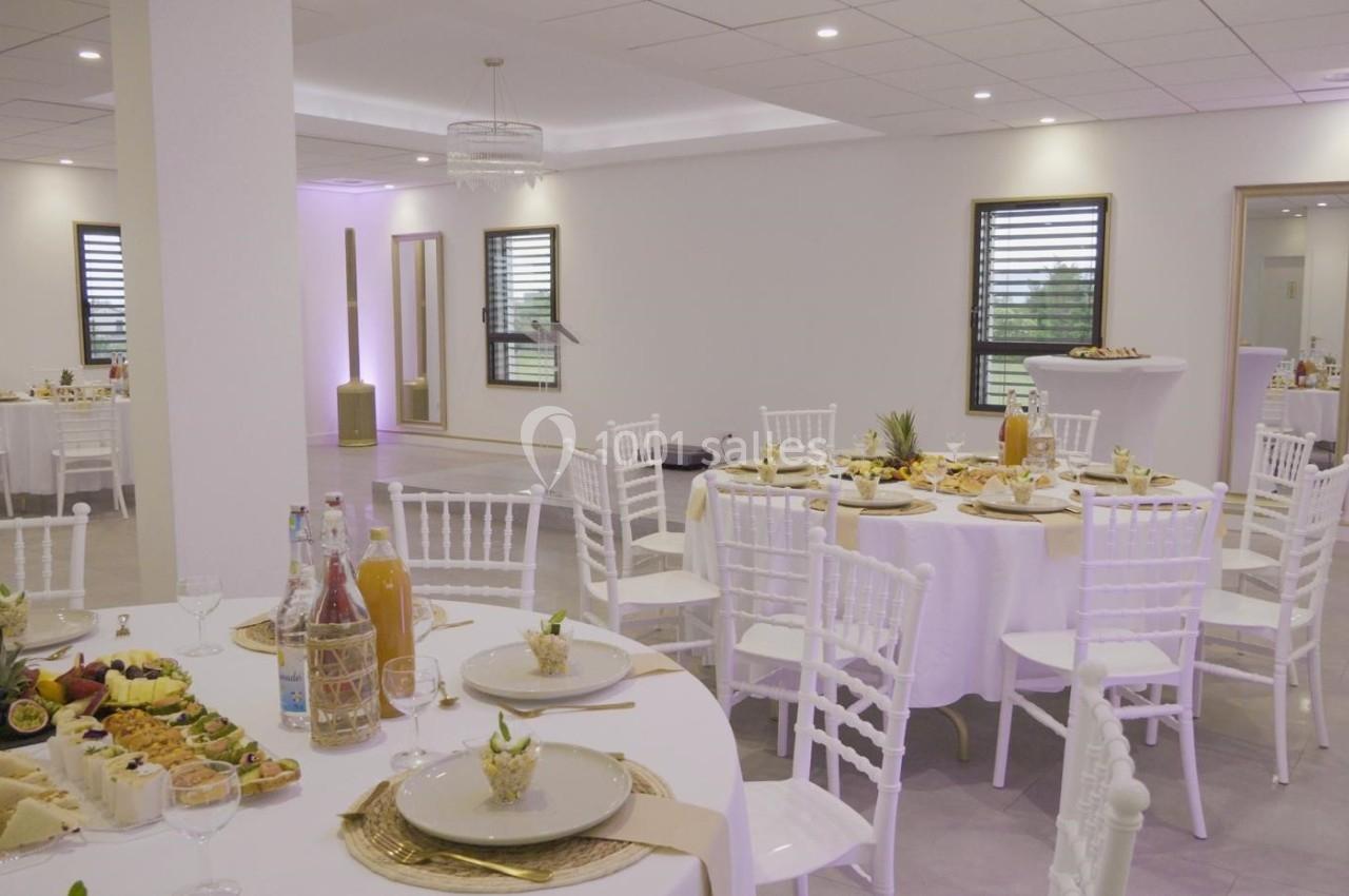 Salle de réception lumineuse avec tables rondes dressées, chaises blanches et buffet de plats variés.