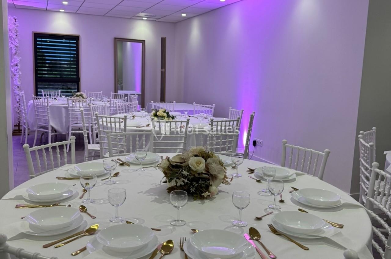 Salle de réception avec tables rondes dressées, nappes blanches, couverts dorés et éclairage violet tamisé.