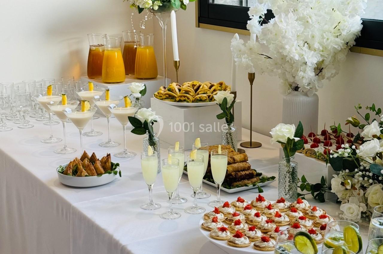 Table dressée avec des boissons, verrines, pâtisseries et fleurs blanches, près d'une fenêtre éclairée.