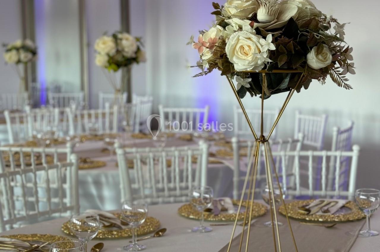 Salle de réception élégante avec tables dressées, centres de table fleuris sur supports dorés et éclairage tamisé.