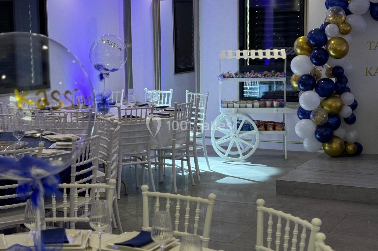 Salle de réception décorée avec des tables dressées, des ballons colorés et un chariot blanc en arrière-plan.
