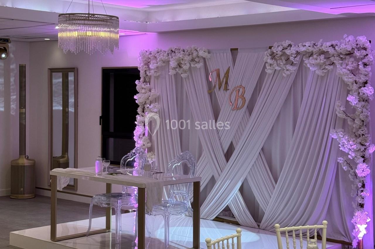 Salle de réception décorée avec des rideaux blancs, des fleurs, des chaises blanches et un éclairage rose.