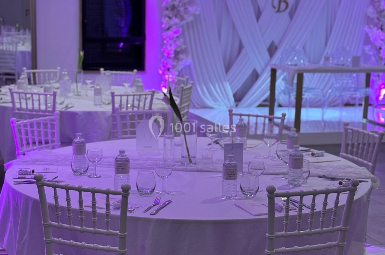Salle de réception décorée avec des tables rondes blanches, des chaises élégantes et un fond lumineux violet.