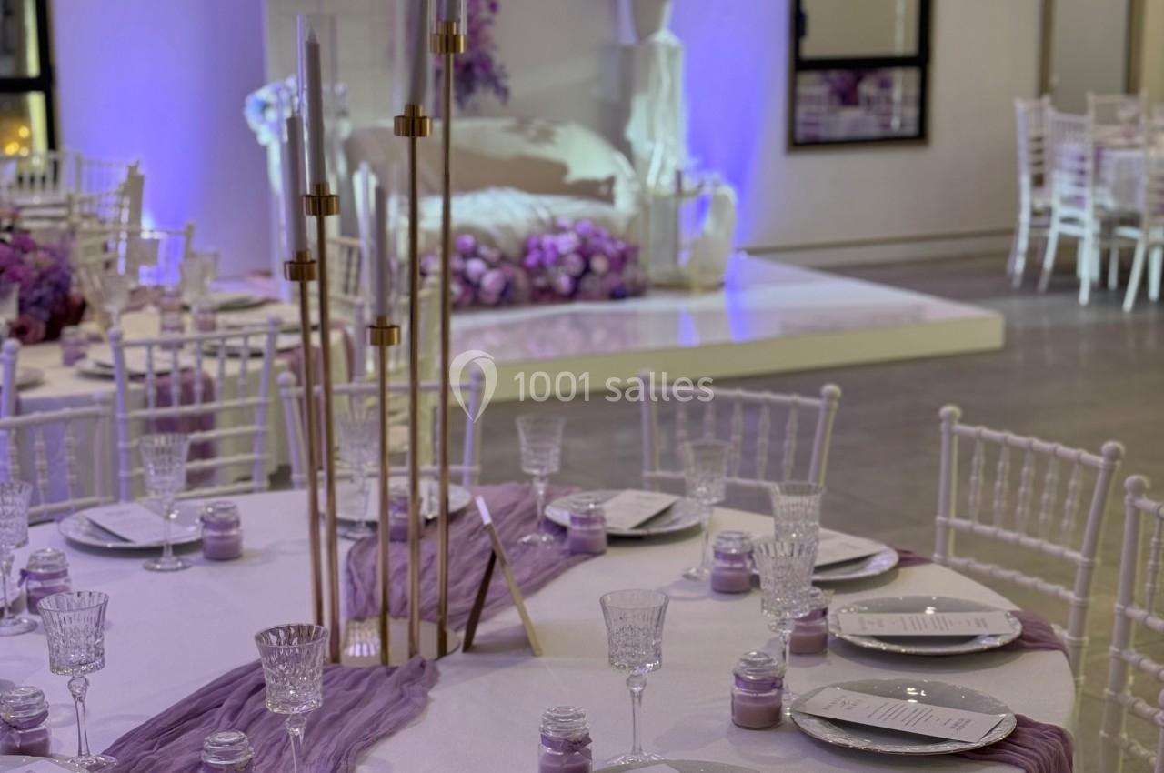 Salle de réception décorée en violet avec tables dressées, chaises blanches et éclairage tamisé.
