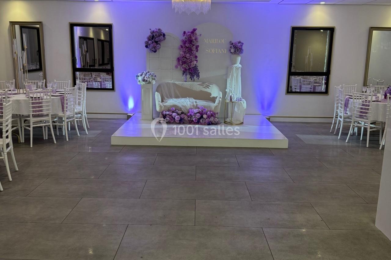 Salle de réception décorée avec des fleurs violettes, un canapé sur une estrade et des tables dressées autour.