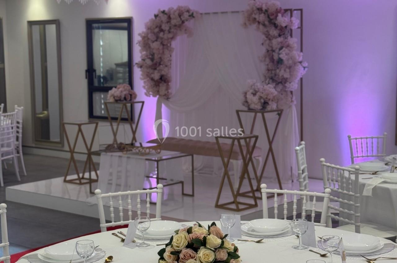 Salle de réception décorée avec des tables dressées, un fond floral rose et blanc, et un éclairage violet.
