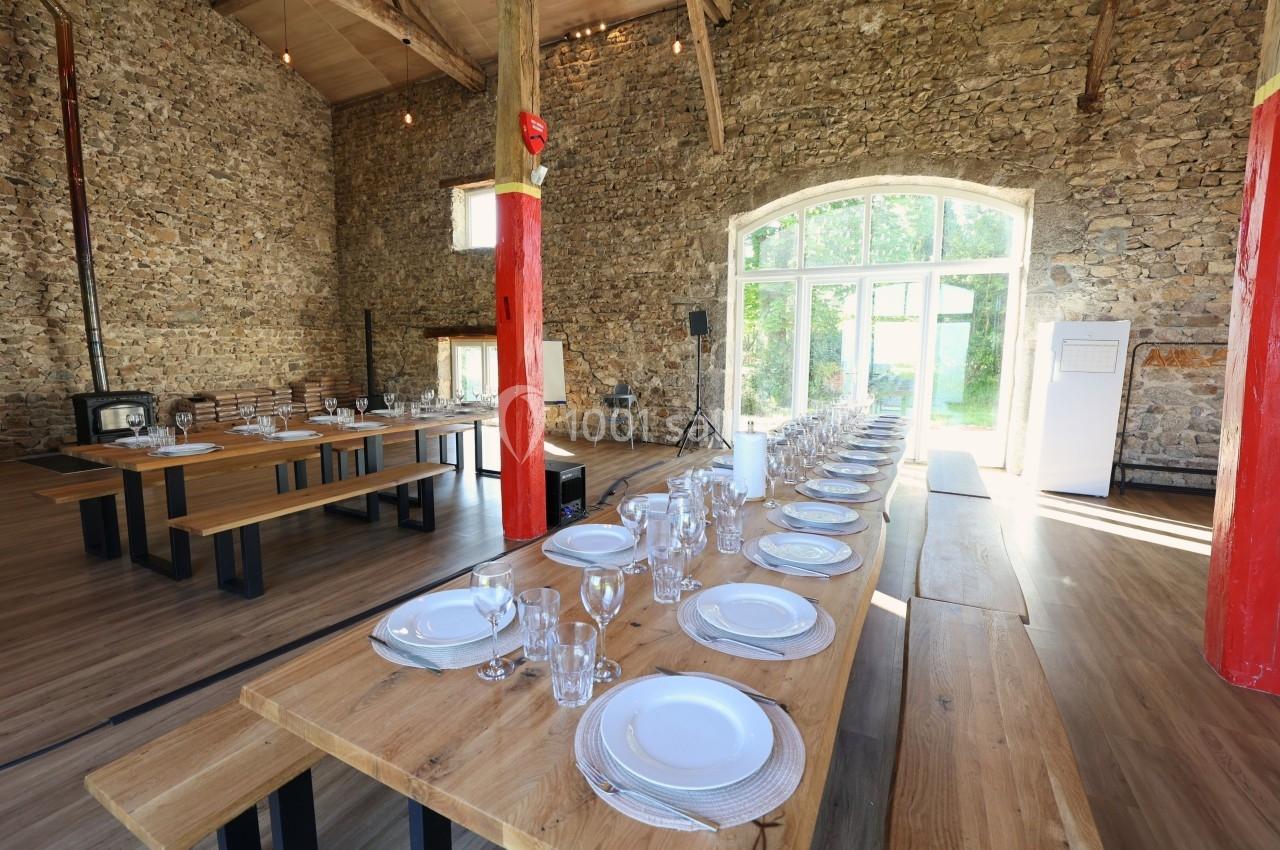 Grande salle en pierre avec de longues tables en bois dressées pour un repas, éclairée par une grande baie vitrée.