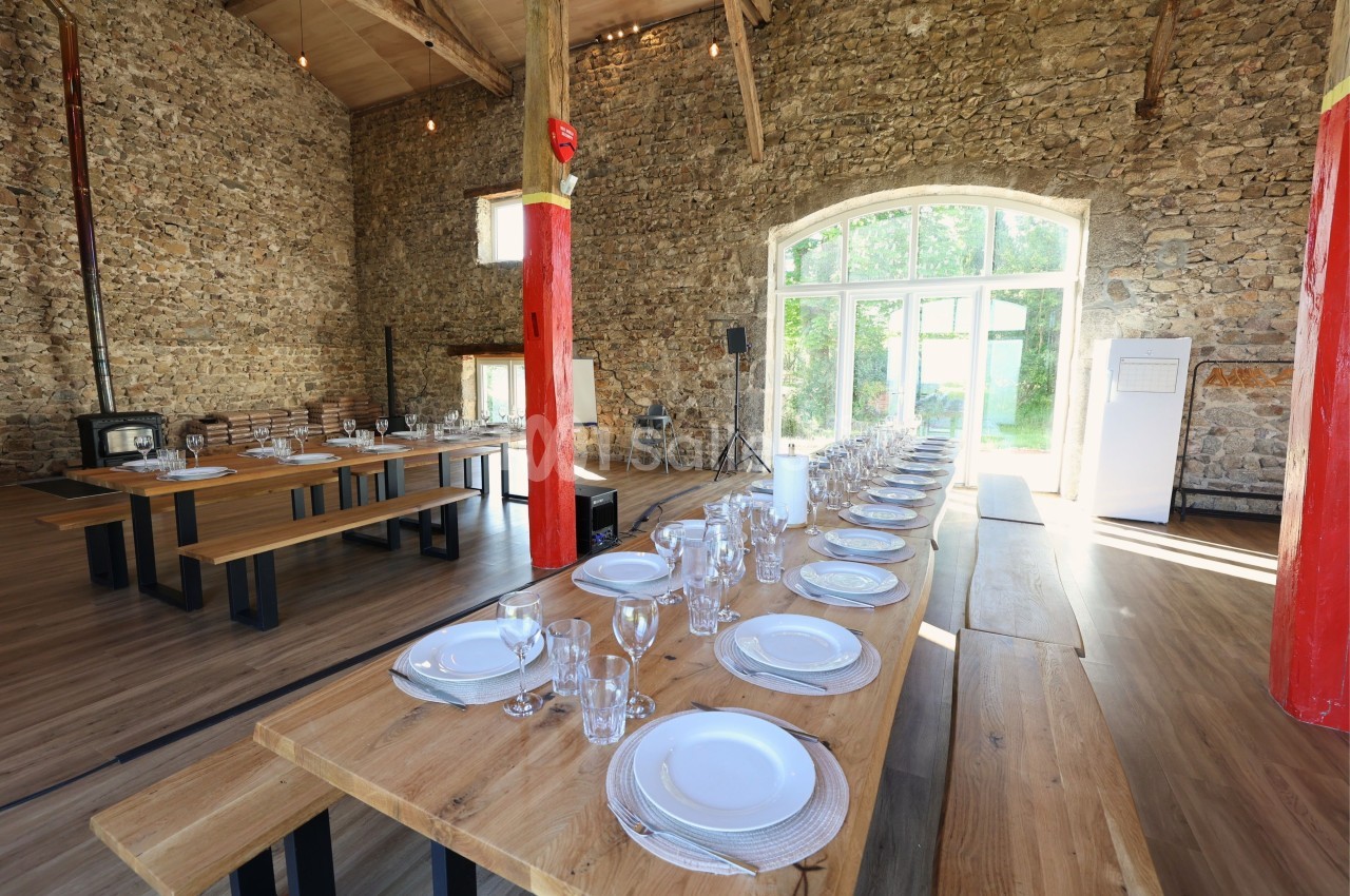 Grande salle en pierre avec de longues tables en bois dressées pour un repas, éclairée par une grande baie vitrée.