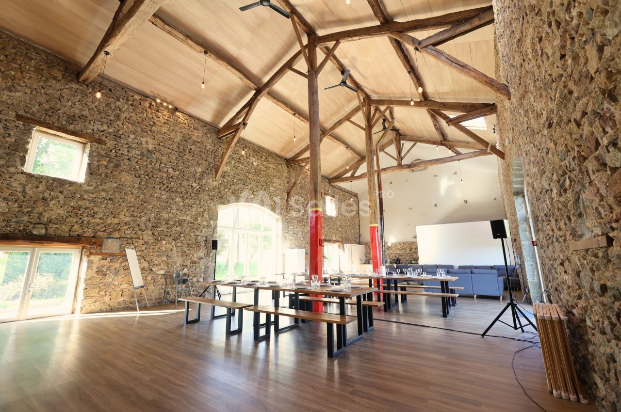 Salle spacieuse avec murs en pierre, poutres apparentes, grandes fenêtres et tables disposées pour un événement.
