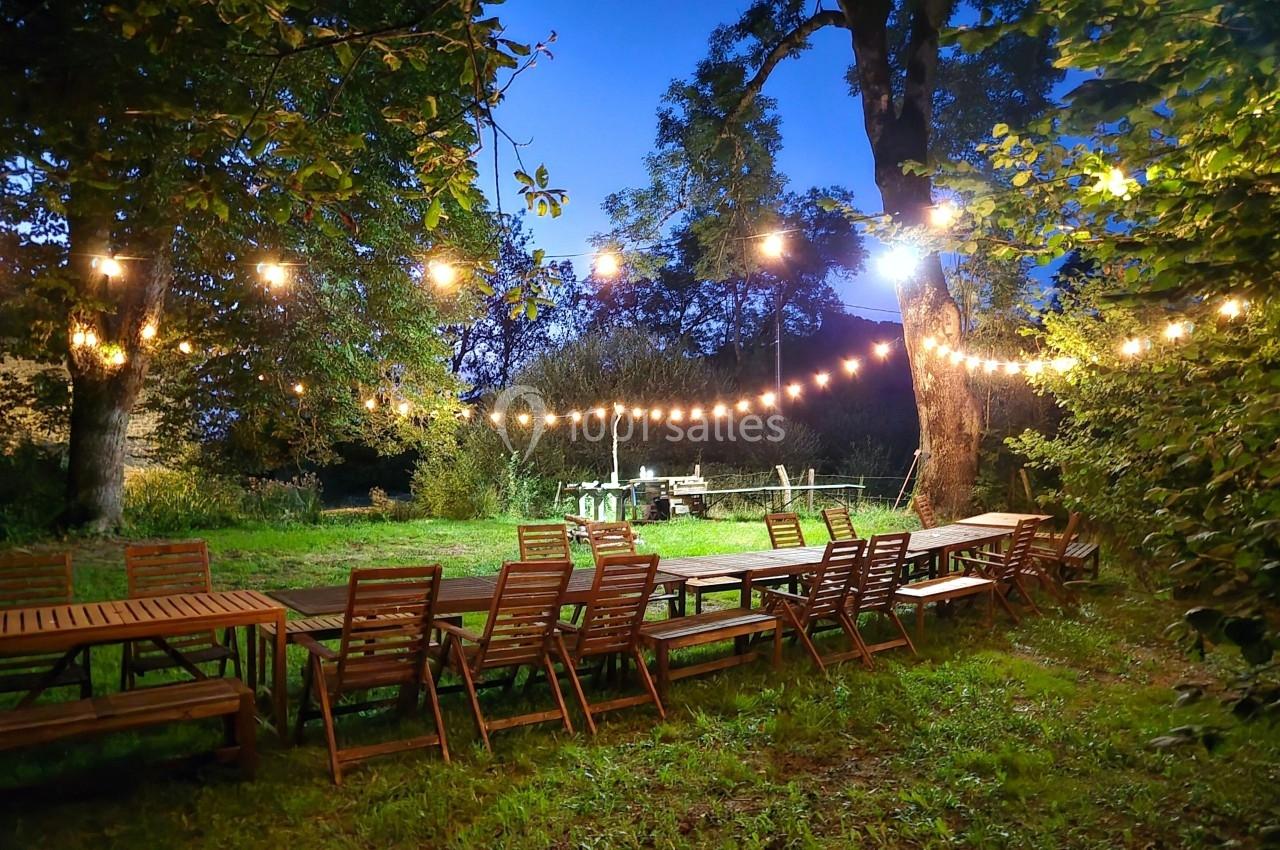 Tables et chaises en bois disposées en extérieur sous des guirlandes lumineuses, entourées d'arbres au crépuscule.