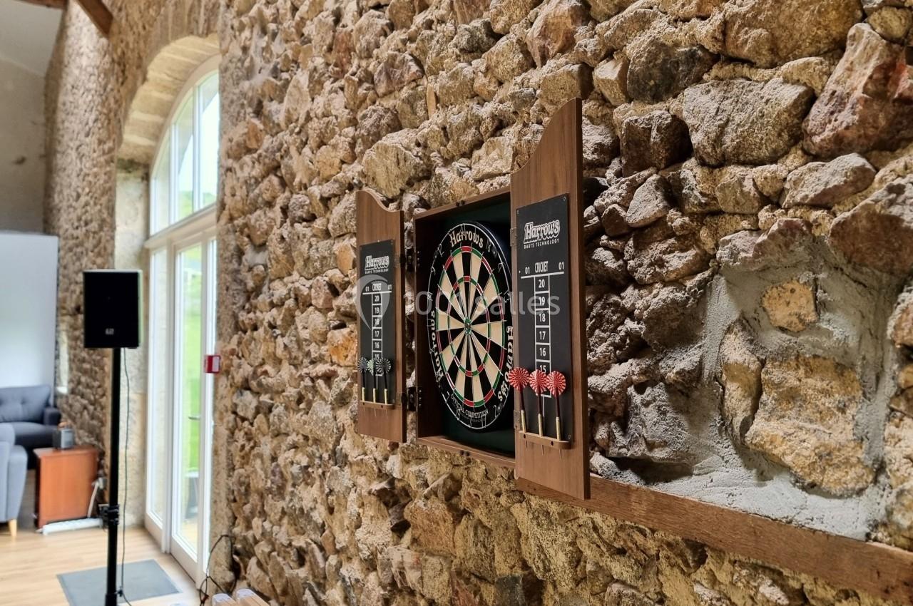 Cible de fléchettes accrochée à un mur en pierre dans une pièce lumineuse avec une grande fenêtre.