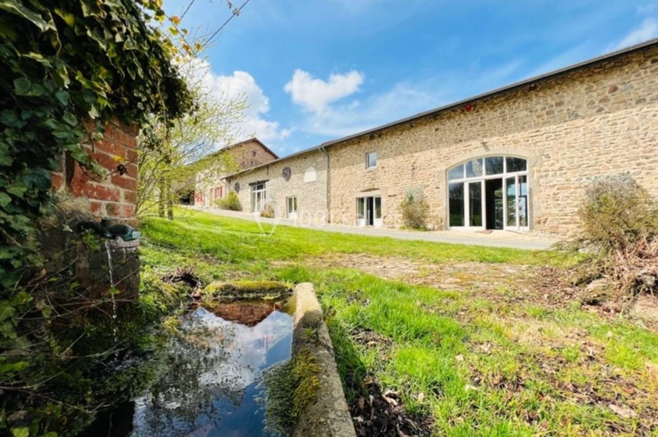 Bâtiment en pierre avec grandes baies vitrées, entouré de verdure, près d'un petit cours d'eau sous un ciel bleu.