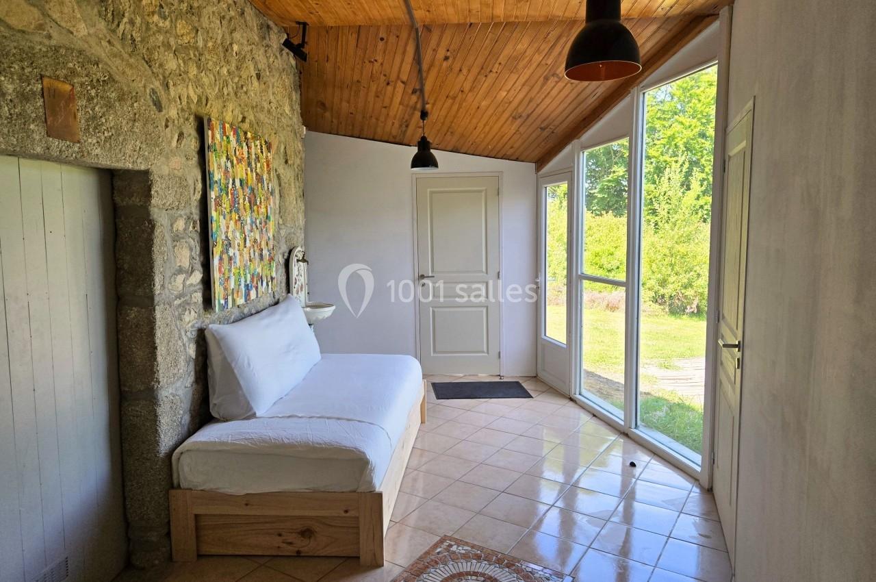 Lit simple près d'un mur en pierre dans une pièce lumineuse avec plafond en bois et grande baie vitrée donnant sur un jardin.