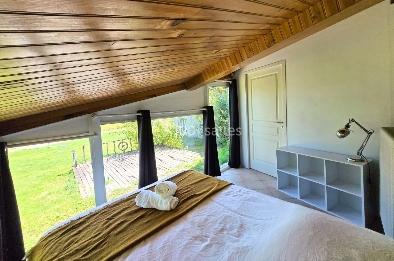 Chambre lumineuse sous combles avec lit double, étagère, lampe de bureau et vue sur un jardin par de grandes fenêtres.