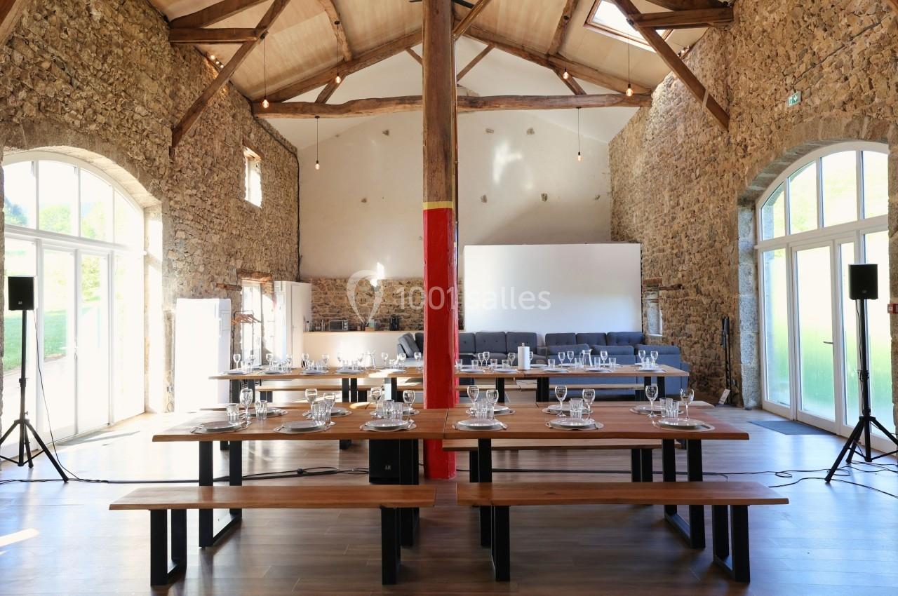 Salle en pierre avec poutres apparentes, tables en bois dressées, grandes baies vitrées laissant entrer la lumière naturelle.