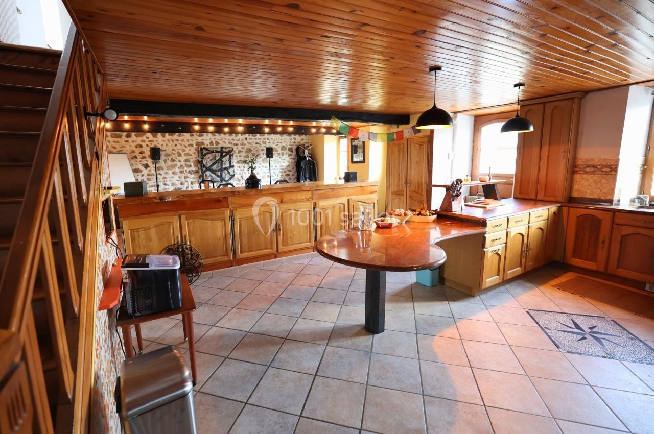 Cuisine rustique avec plafond en bois, meubles en bois clair, table ronde centrale et éclairage suspendu.