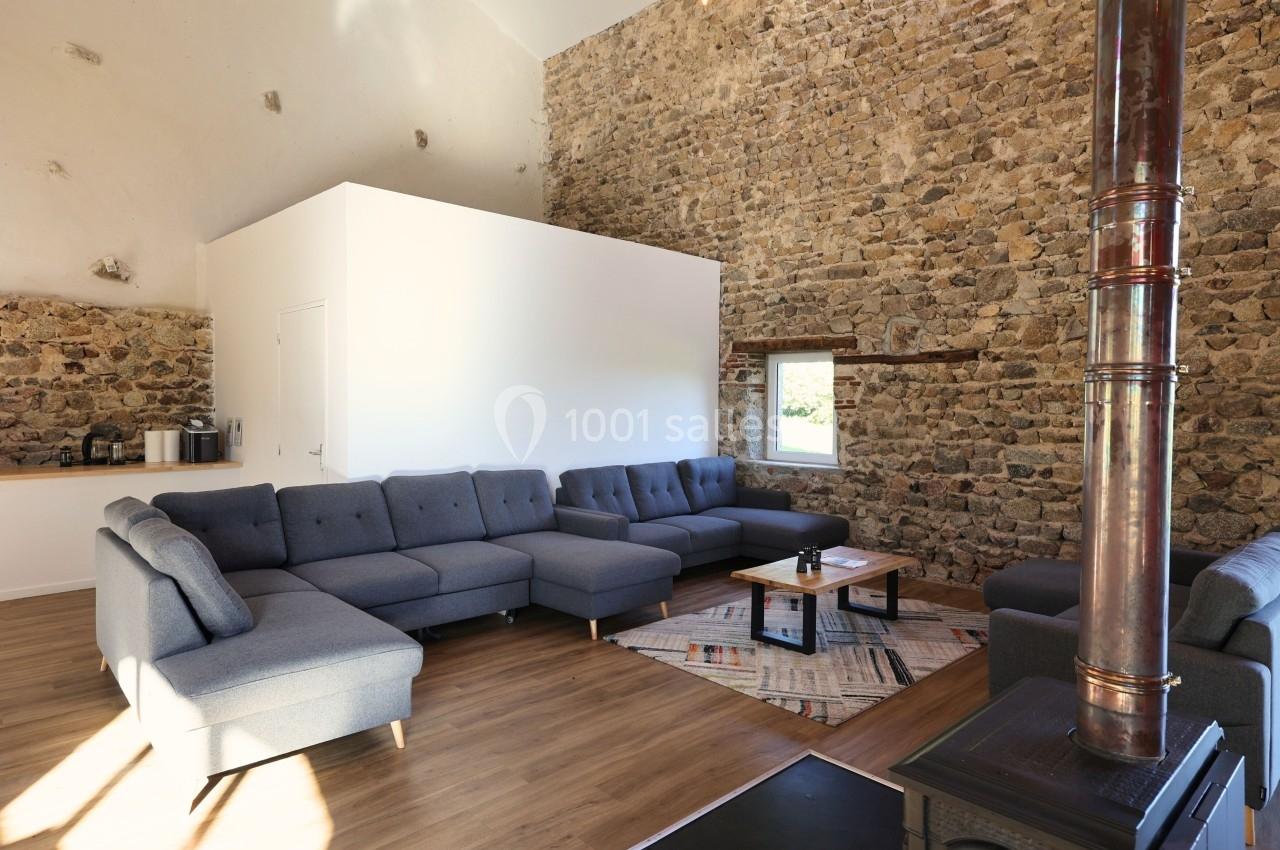 Salon spacieux avec murs en pierre, canapé d'angle gris, poêle à bois et lumière naturelle provenant d'une fenêtre.