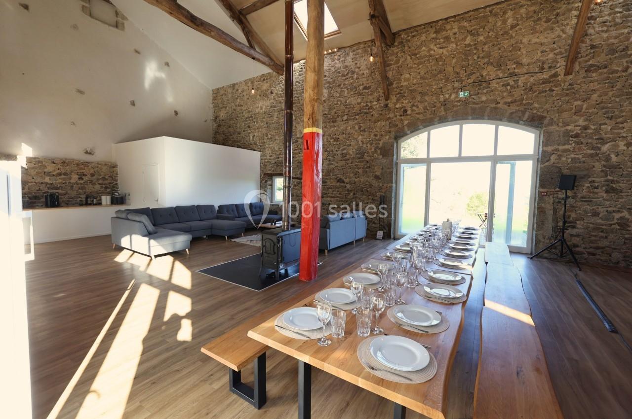 Grande salle lumineuse avec murs en pierre, table en bois dressée pour un repas et canapés dans un coin salon.