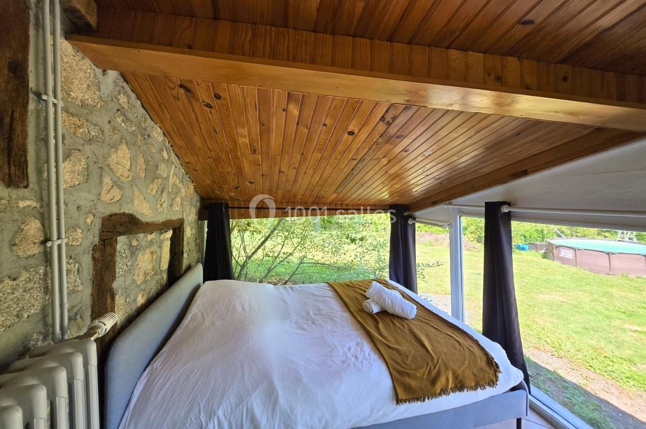 Lit double avec draps blancs et couverture ocre, situé dans une chambre avec plafond en bois et vue sur un jardin verdoyant.