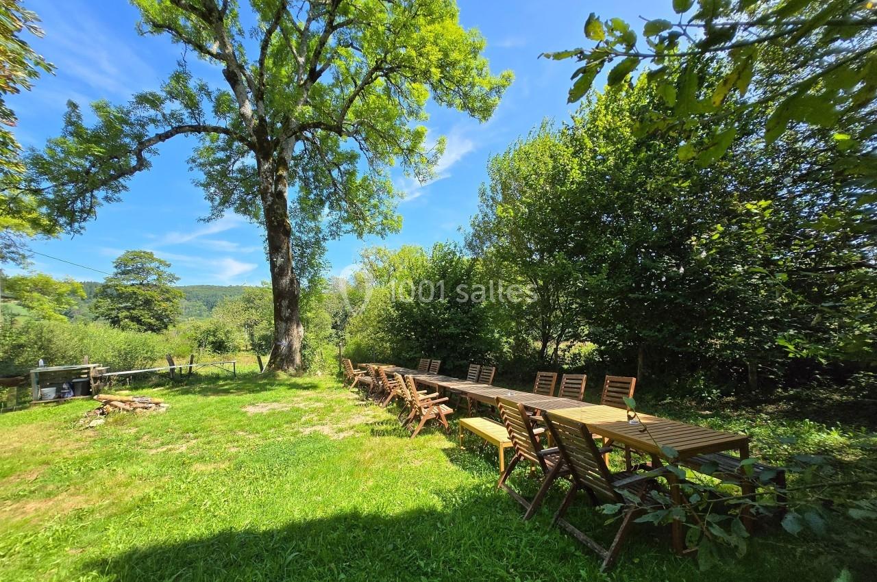 Grande table en bois entourée de chaises dans un jardin verdoyant, sous un arbre imposant par une journée ensoleillée.