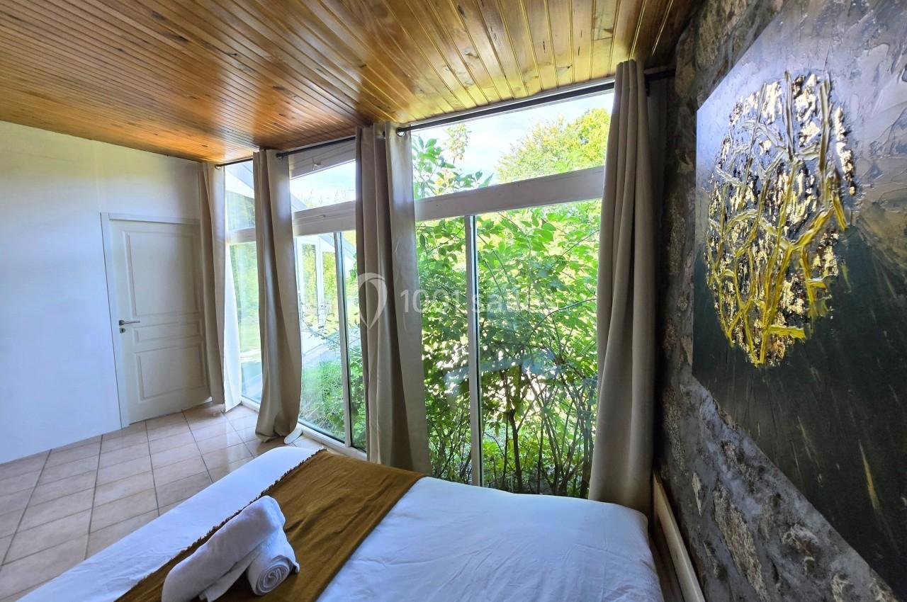 Chambre lumineuse avec grande baie vitrée donnant sur un jardin verdoyant, plafond en bois et lit avec linge plié.