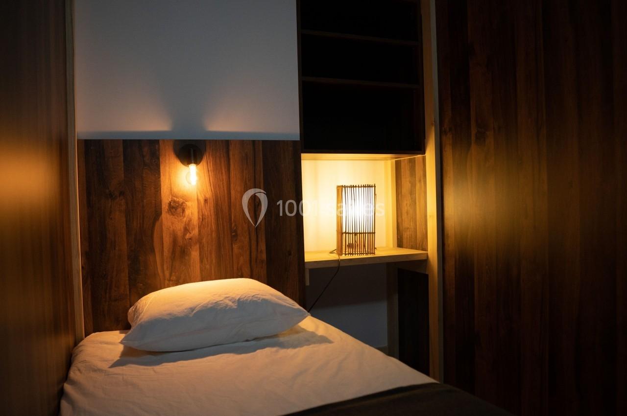 Lit simple avec oreiller et lampe allumée sur une table de chevet, dans une chambre aux murs en bois sombre.