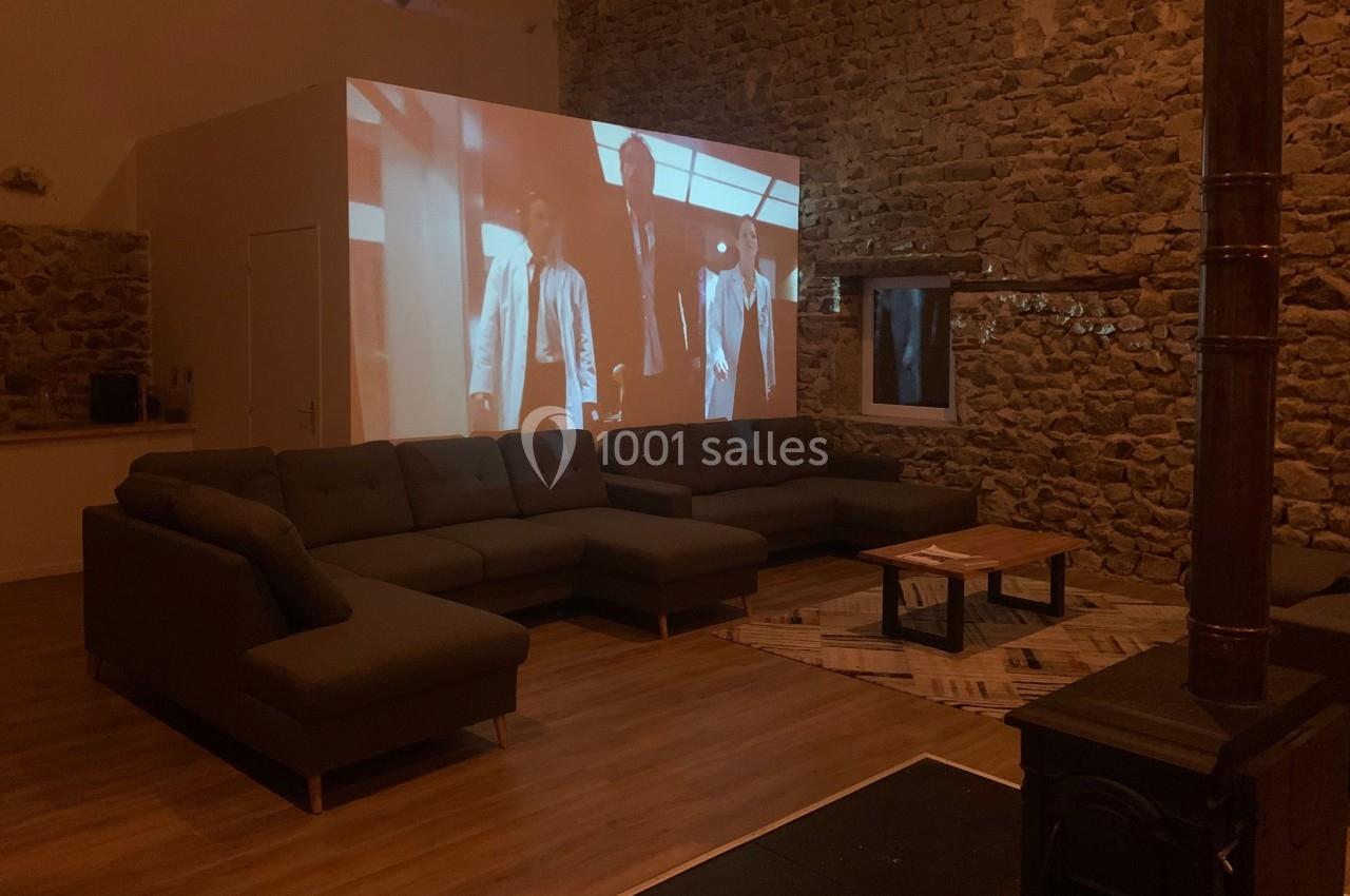 Projection d'un film sur un mur en pierre dans un salon avec canapé d'angle, table basse et poêle à bois.