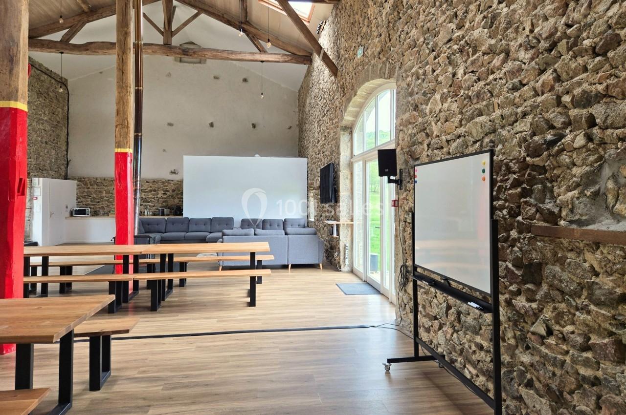 Salle lumineuse avec murs en pierre, grandes fenêtres, tables en bois, tableau blanc et canapés gris au fond.