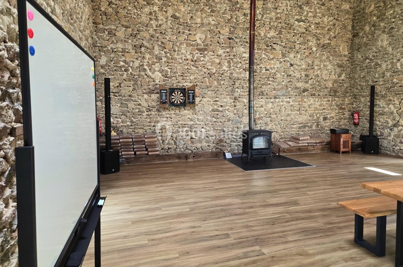 Salle avec murs en pierre, tableau blanc, poêle à bois, cible de fléchettes et bancs en bois sur un sol en parquet.