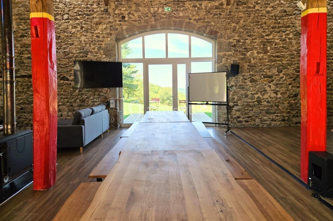 Salle en pierre avec une grande baie vitrée, table en bois, canapé, écran et tableau blanc, vue sur l'extérieur verdoyant.