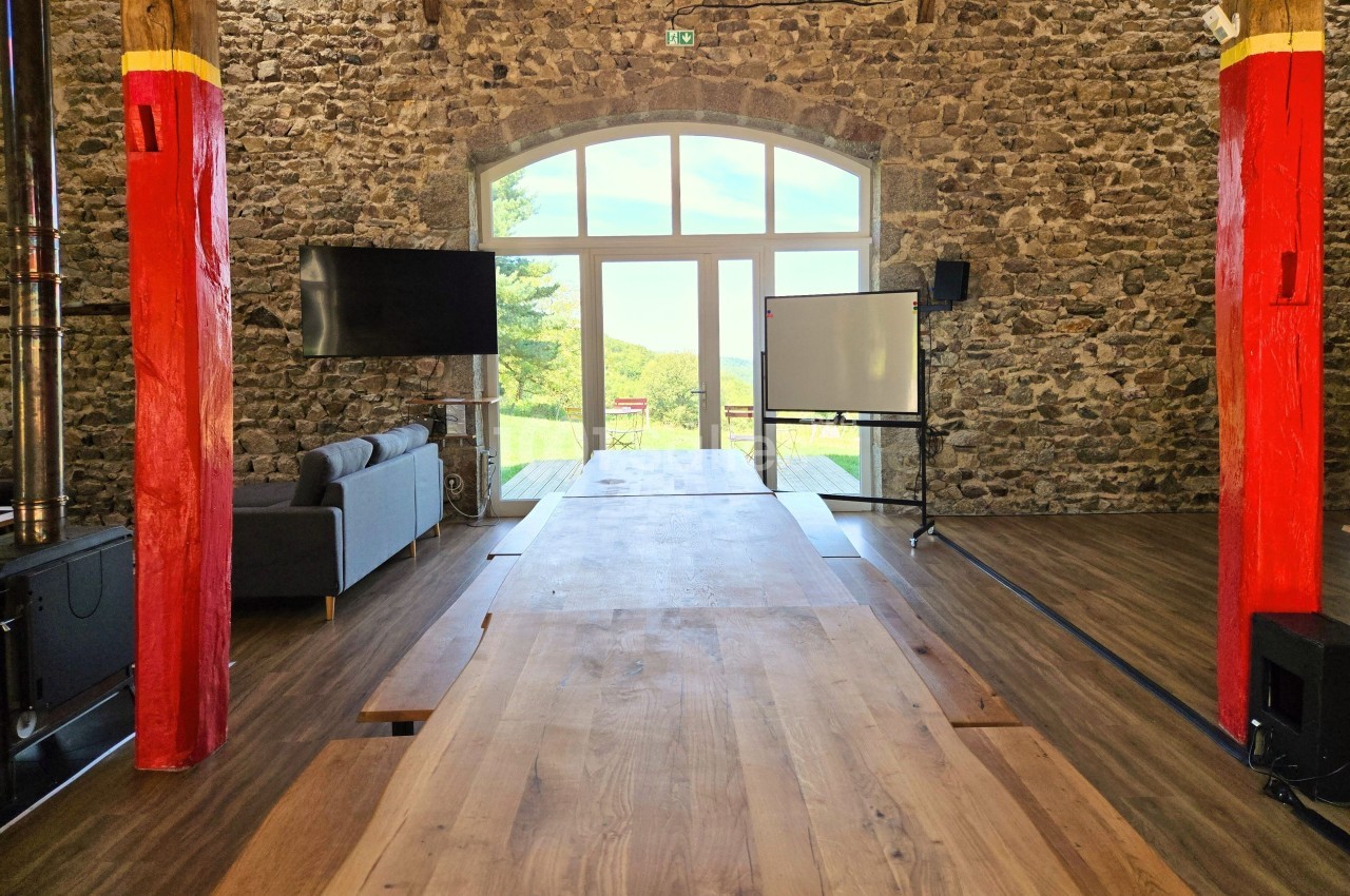 Salle en pierre avec une grande baie vitrée, table en bois, canapé, écran et tableau blanc, vue sur l'extérieur verdoyant.