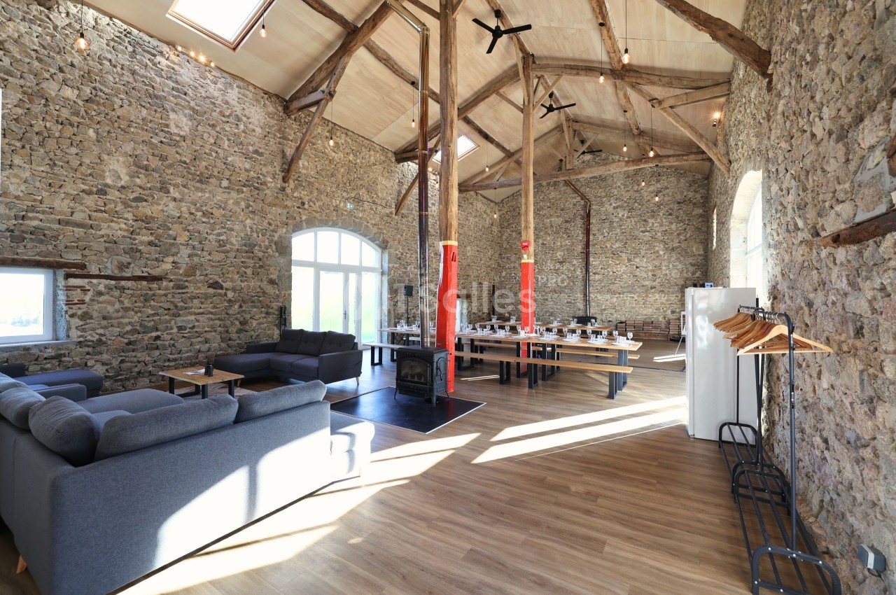 Salle spacieuse avec murs en pierre, poutres apparentes, canapés, tables dressées et lumière naturelle abondante.