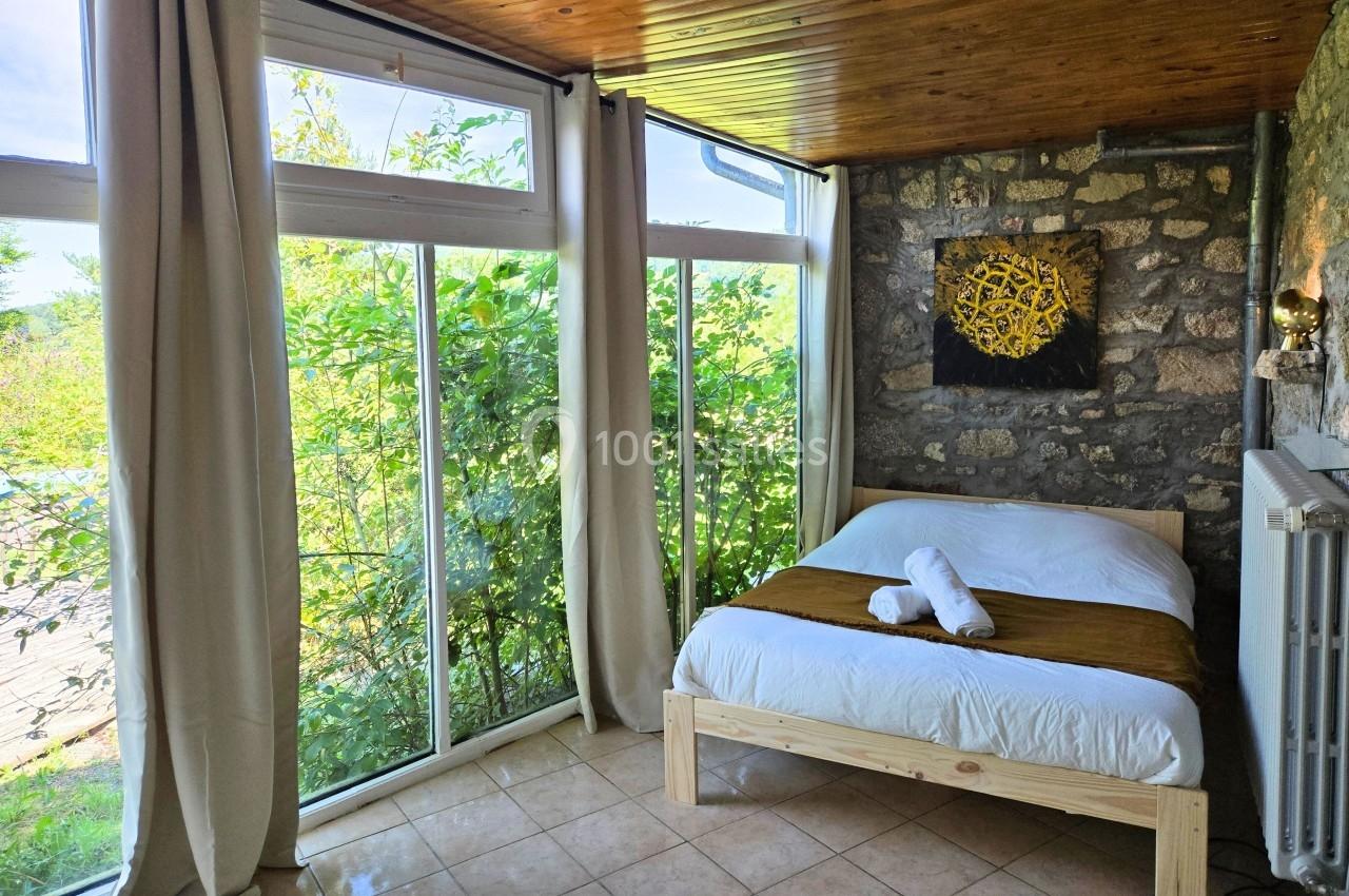 Chambre lumineuse avec lit double, grandes baies vitrées donnant sur un jardin verdoyant et mur en pierre apparente.