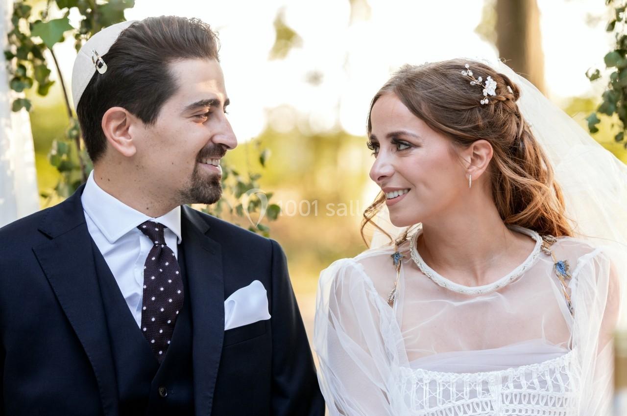 Un couple en tenue de mariage se regarde avec le sourire, en extérieur sous une lumière naturelle.