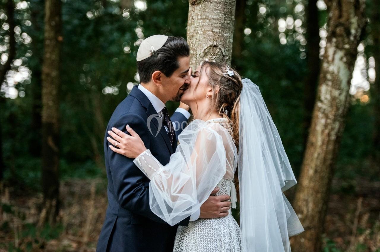 Un couple en tenue de mariage s'embrasse tendrement devant un arbre dans une forêt.