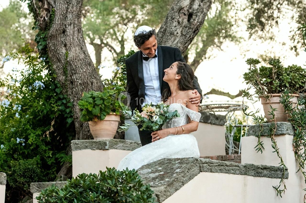 Un couple en tenue de mariage, assis sur des marches en pierre entourées de plantes et d'arbres.