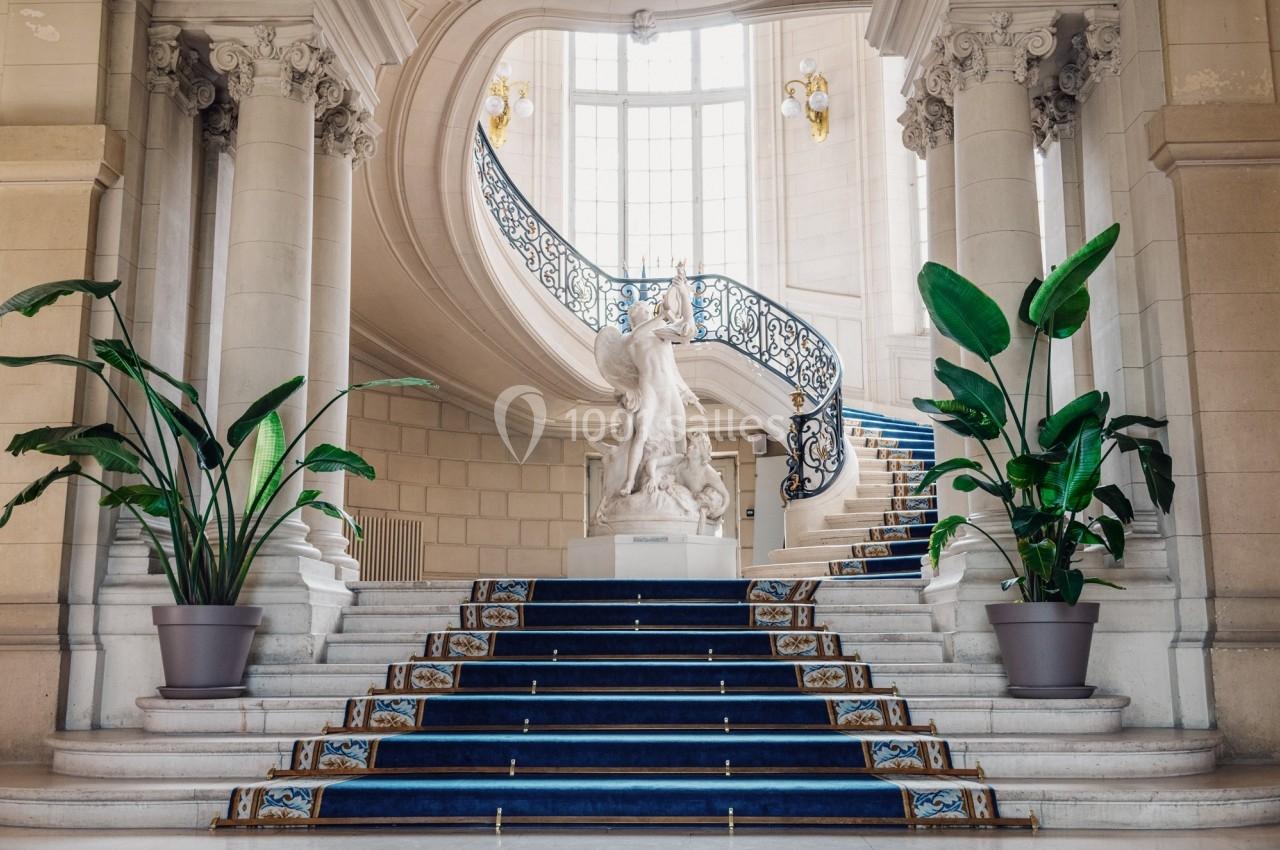 Escalier monumental avec tapis bleu, statue centrale, balustrade ornée et plantes en pots dans un intérieur lumineux.