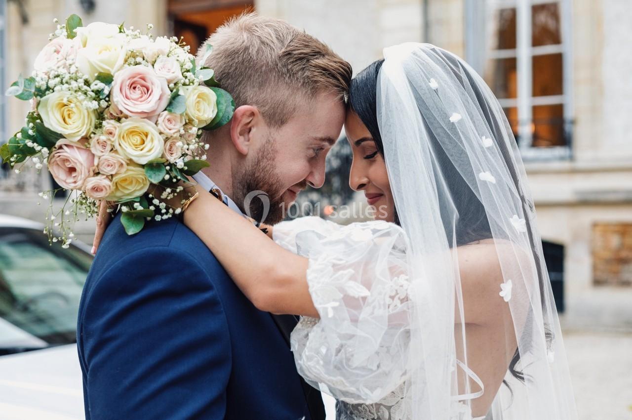 Un couple de mariés s'enlace tendrement, la mariée tenant un bouquet de fleurs pastel.
