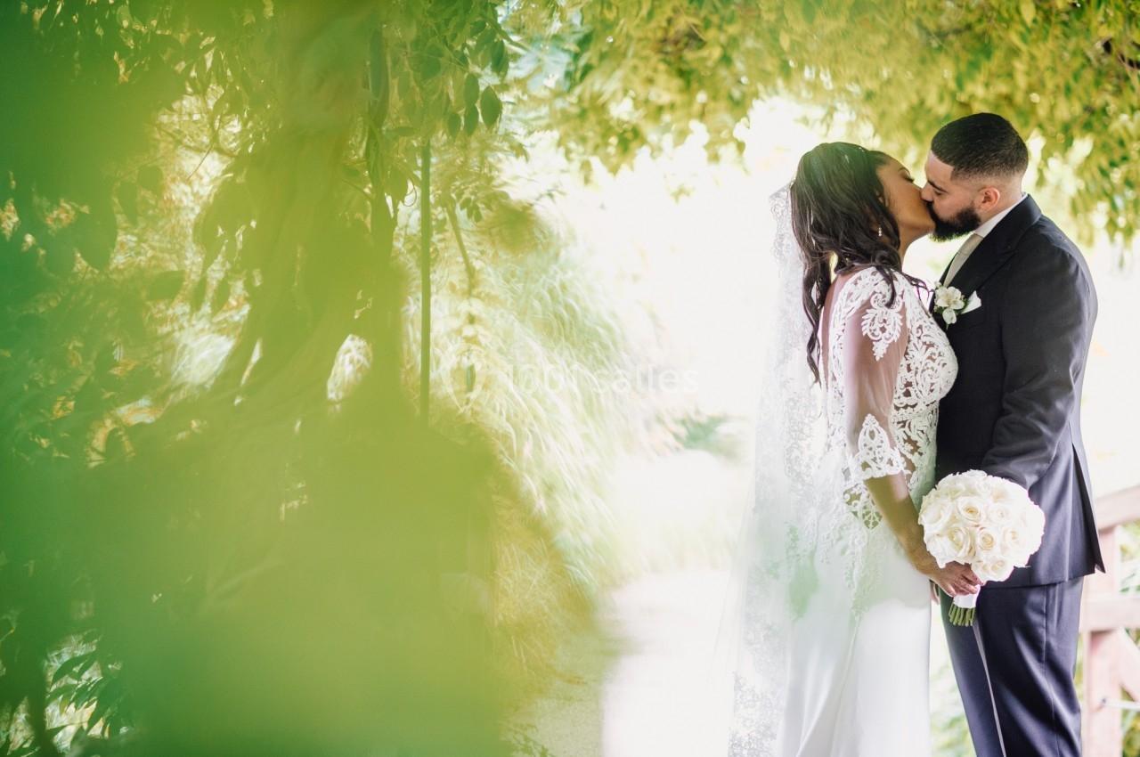 Un couple en tenue de mariage s'embrasse sous une arche de verdure, la mariée tenant un bouquet blanc.